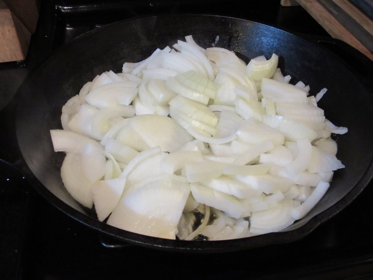 Lots of sliced onions in a cast iron skillet on the stovetop.