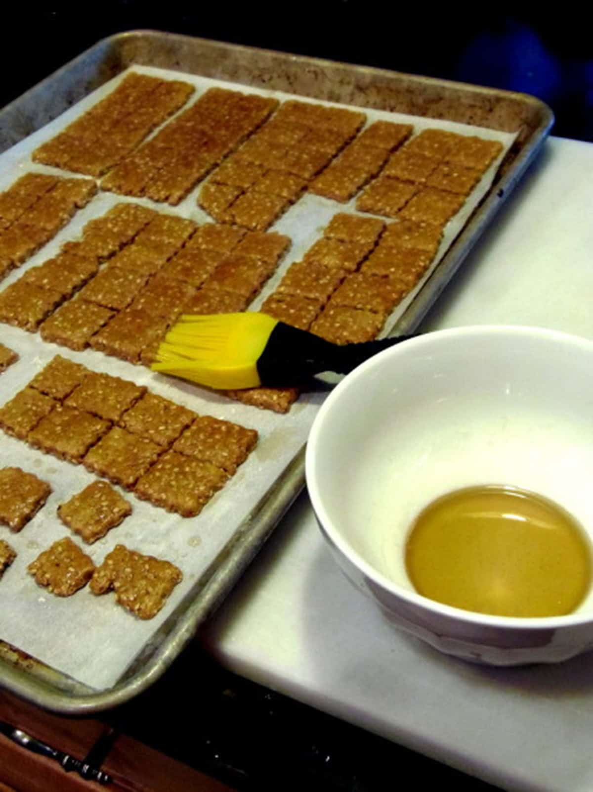 A bowl of honey and a pastry brush next to a baking sheet of homemade sesame seed crackers.