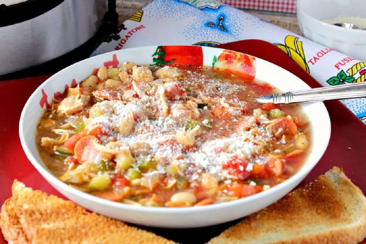 A spoon in a bowl of Italian pepperoni soup with chicken, tomatoes, pepperoni and grated cheese on top.