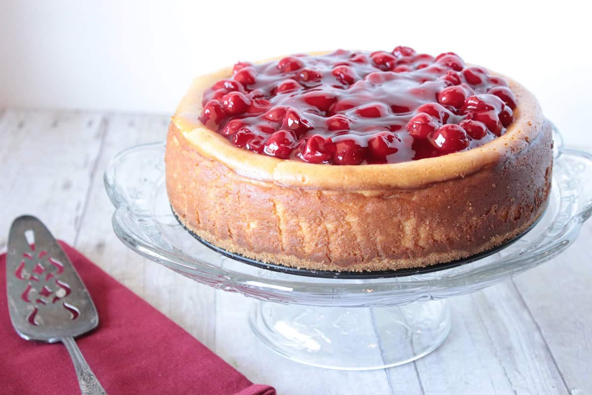 A baked cheesecake with a glistening cherry topping on a glass cake stand.