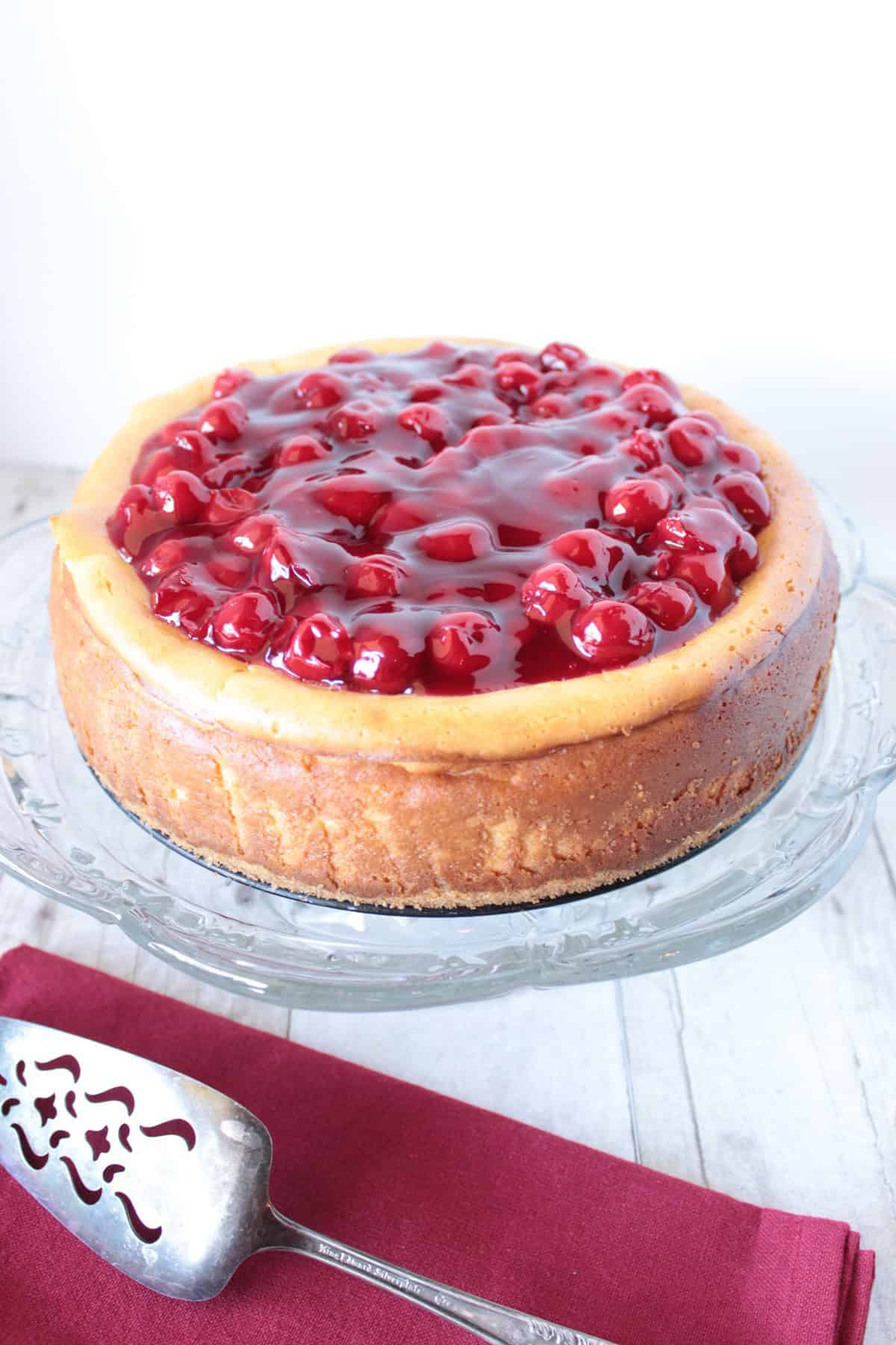 A cherry pie filling topped baked cheesecake with a red napkin and pie server.