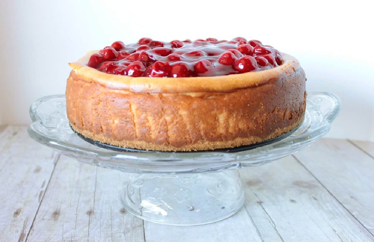 A beautifully baked old-fashioned cheesecake topped with cherry pie filling on a glass cake stand.