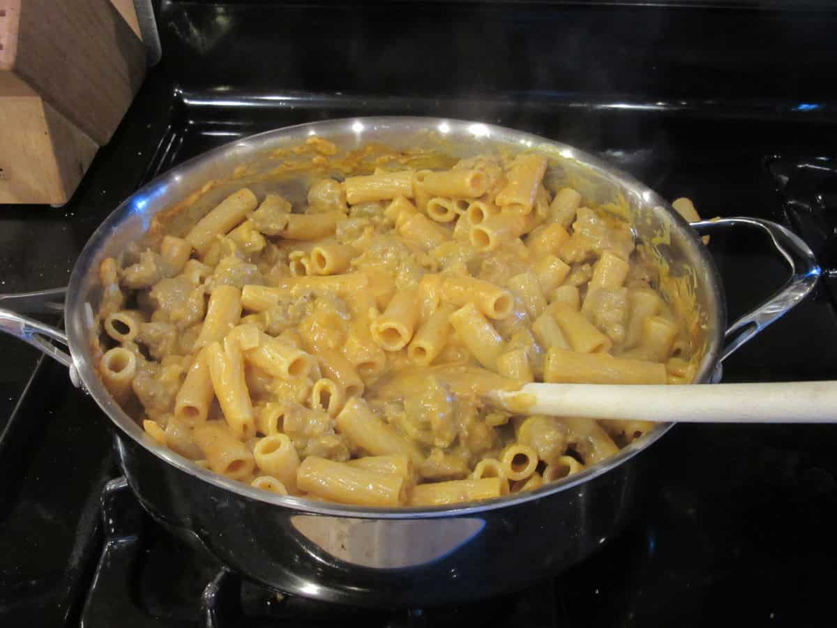 A large skillet on the stovetop filled with cooked rigatoni, bratwurst and pumpkin sauce.