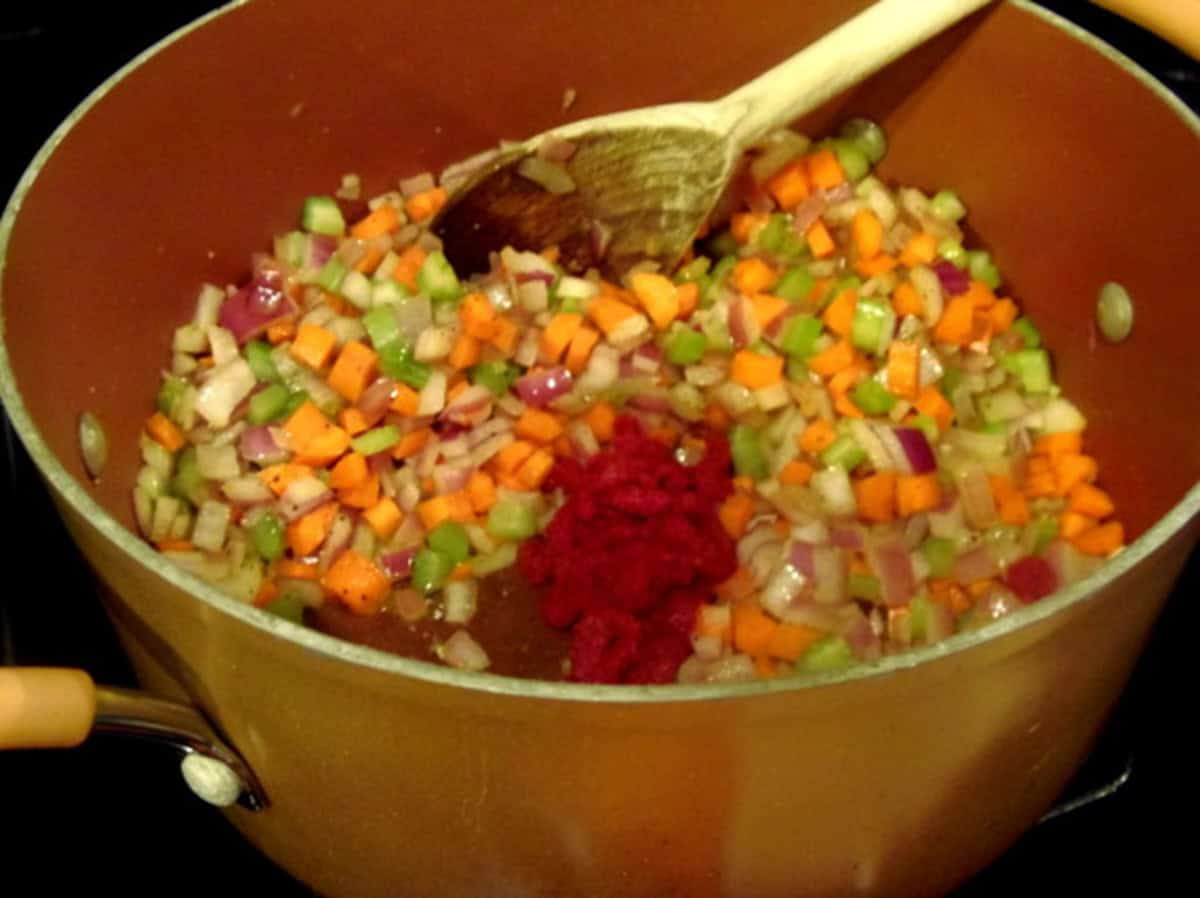 Diced carrots, onions, celery, and tomato paste in a soup pot along with a wooden spoon.