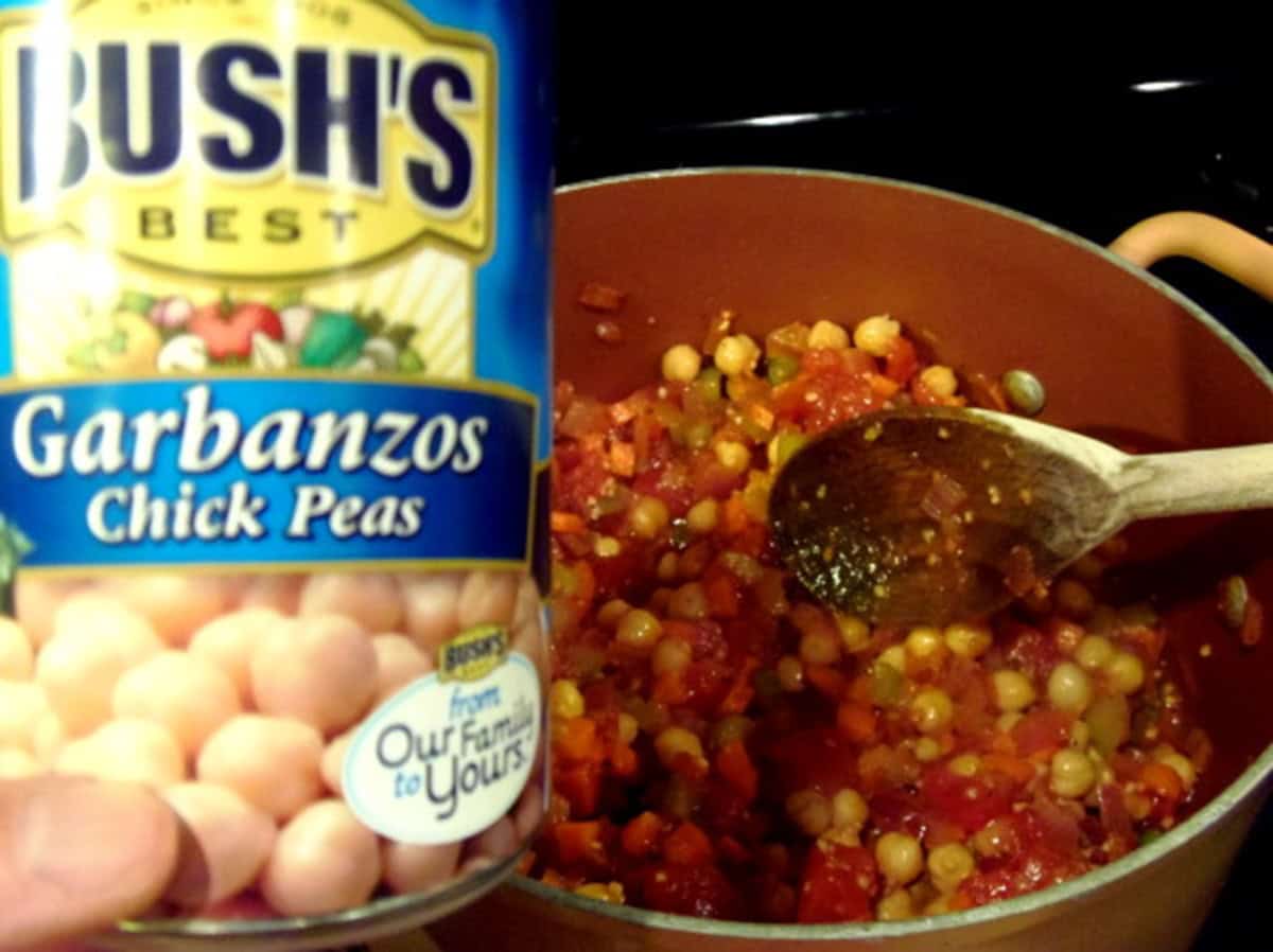 A can of chickpeas being held over a soup pot with the makings of a soup.