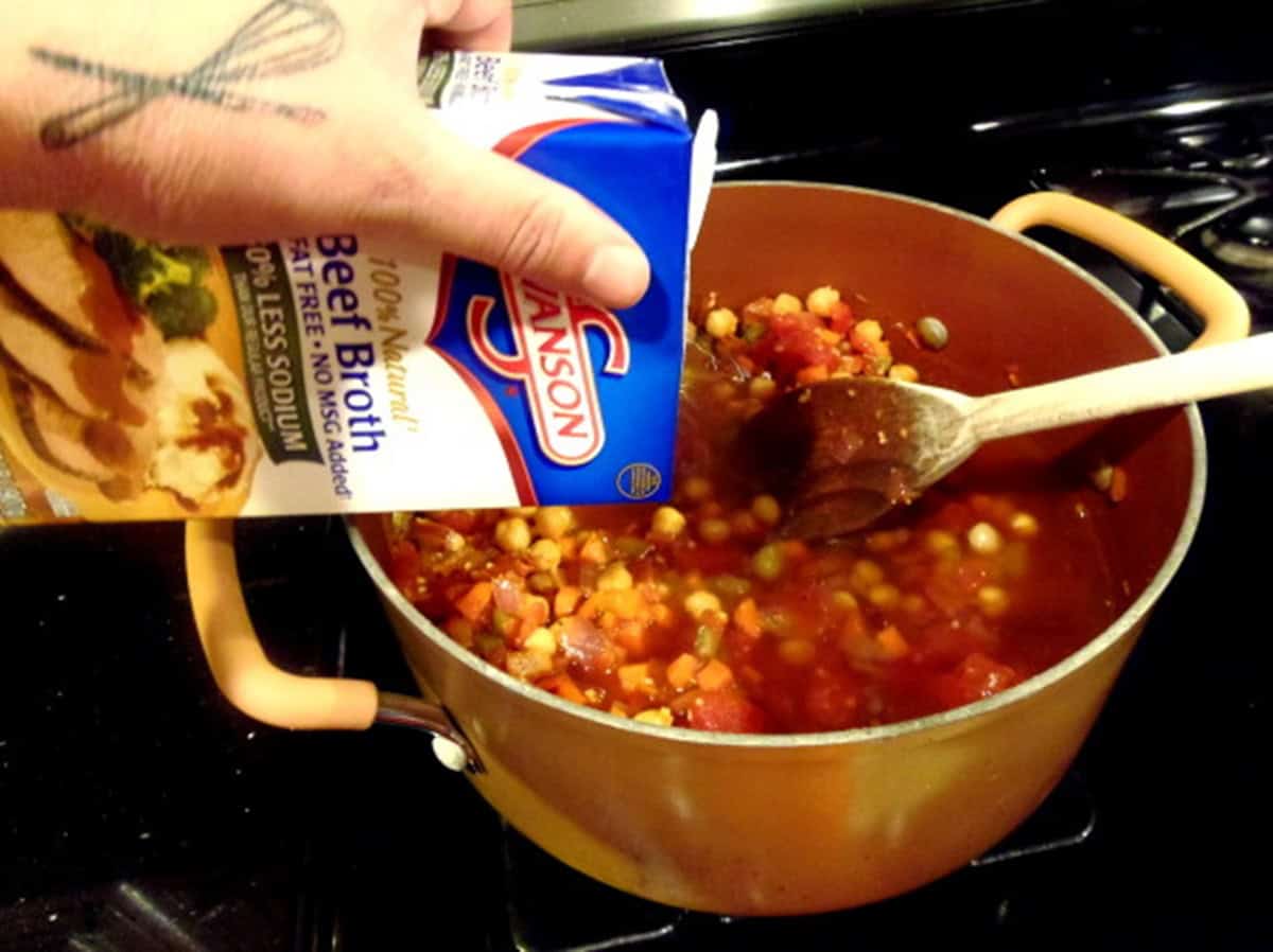 A carton of beef broth being added to a soup pot with chickpeas and veggies.