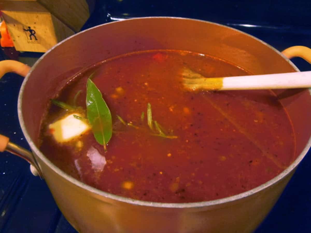 A deep, rich soup broth with a bay leaf, rosemary, and butter on the stovetop.