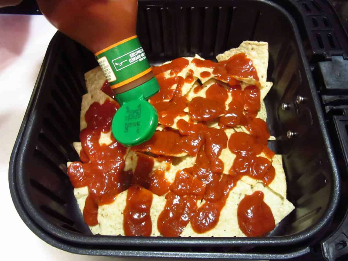 Squeeze pizza sauce being added on top of nacho chips in an air fryer basket.