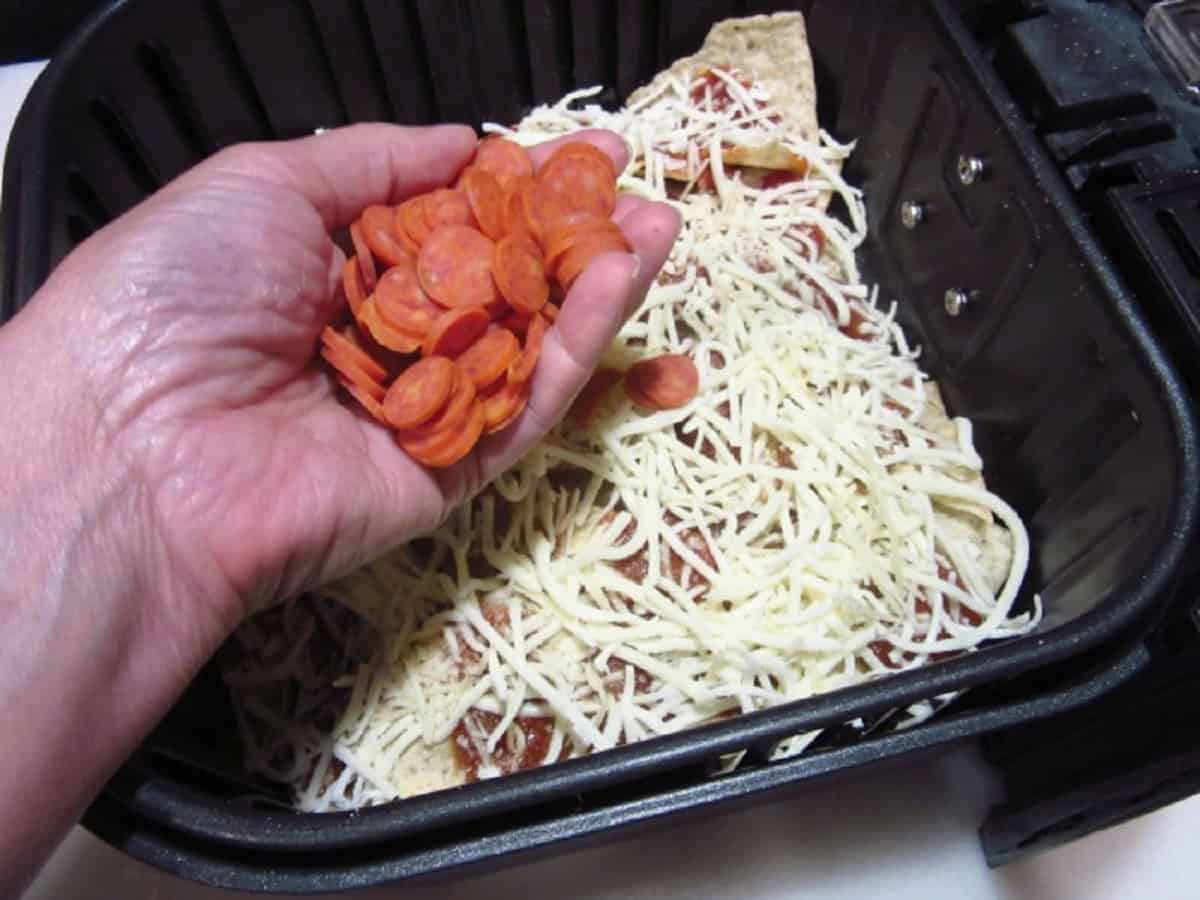 A hand holding a handful of mini pepperoni over an air fryer basket.