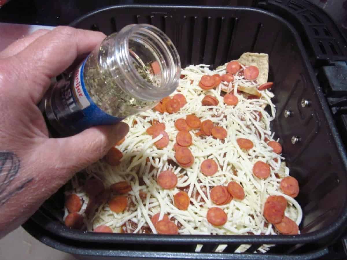 A hand holding a jar or dried basil over an air fryer basket making pizza nachos.