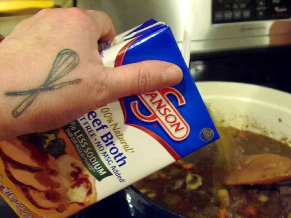 A hand pouring beef broth from a carton into a Dutch oven.