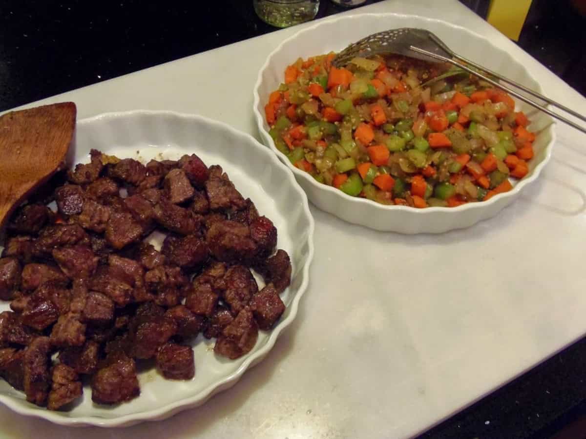 Two shallow dishes on the counter; one with browned beef cubes and one with sauteed veggies.