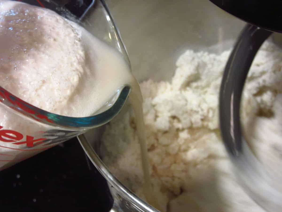 A foamy yeast mixture being added to a stand mixer with flour for making bread.