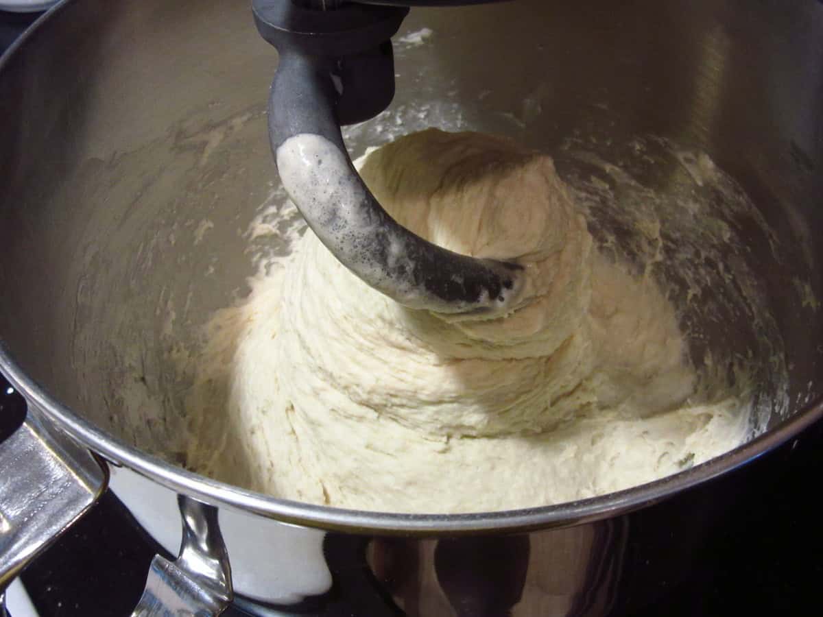 A wet and sticky English muffin bread dough in a stand mixer bowl with a dough hook.