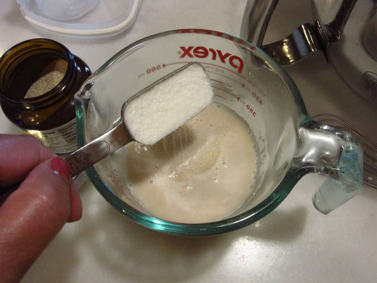 A teaspoon of sugar being added into a glass measuring cup of dissolved yeast and water.