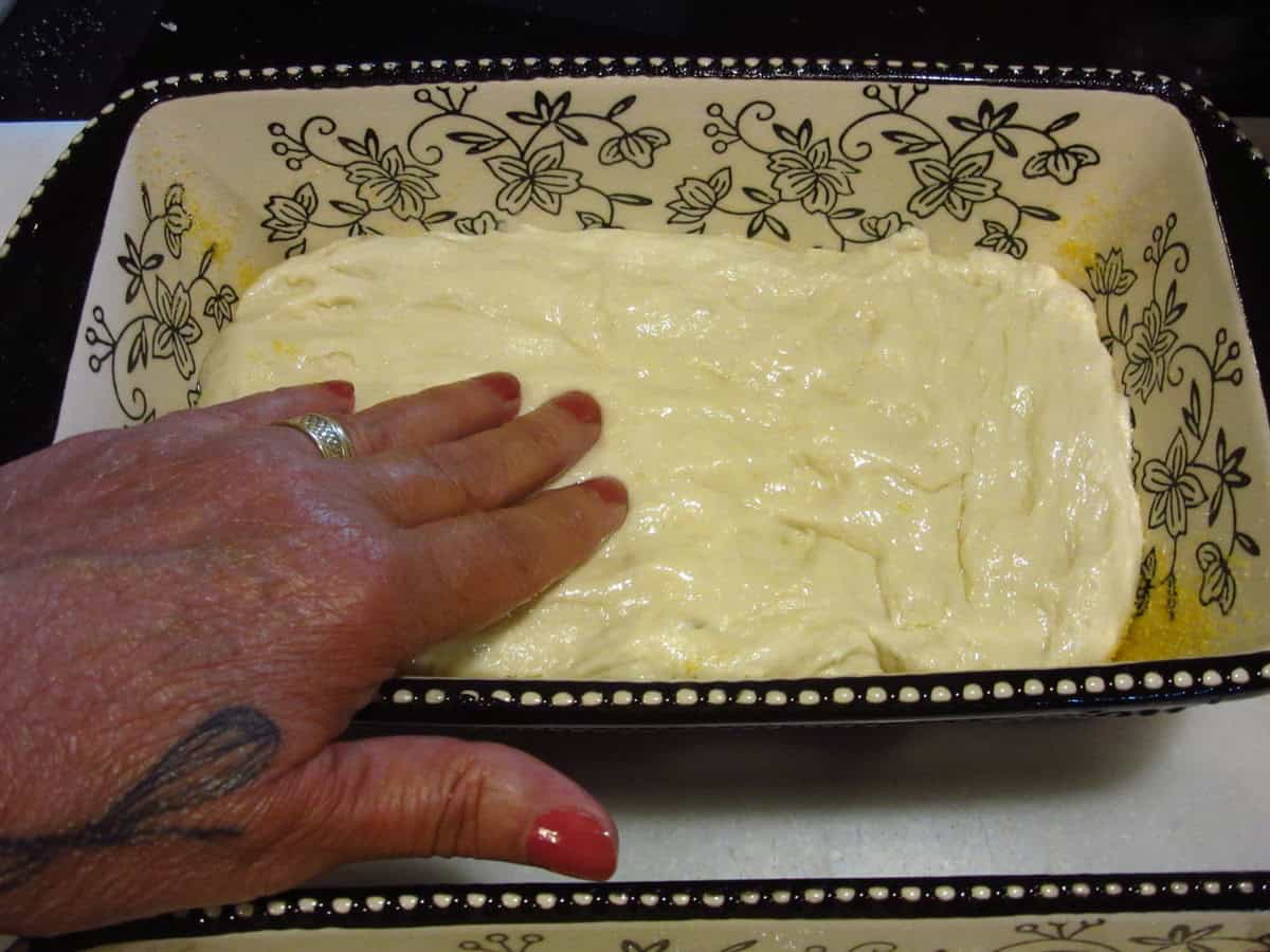 Fingertips pressing an oiled bread dough into a ceramic loaf pan.