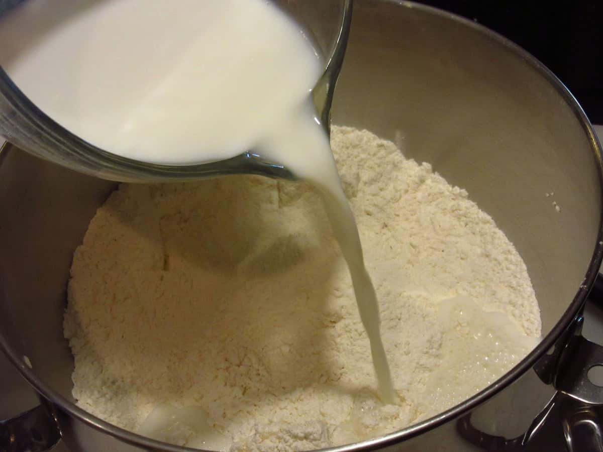 Milk being poured into a stand mixer bowl that has all-purpose flour inside.