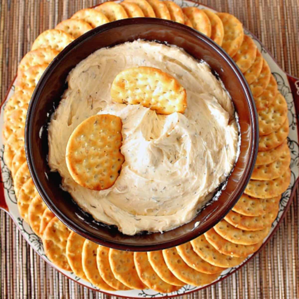 Two crackers stuck in a roasted garlic dip bowl with additional crackers around the bowl.