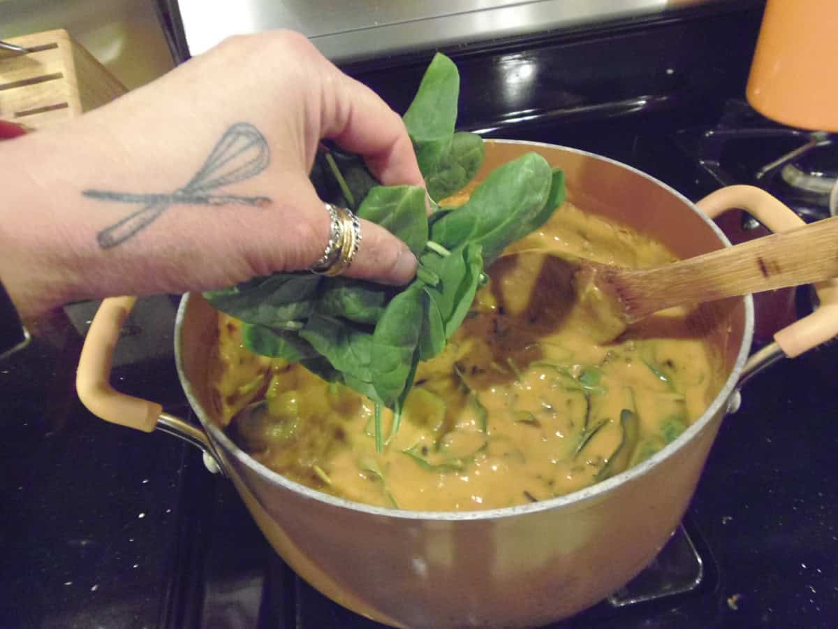 A hand adding a bunch of fresh spinach to a chowder on the stovetop.