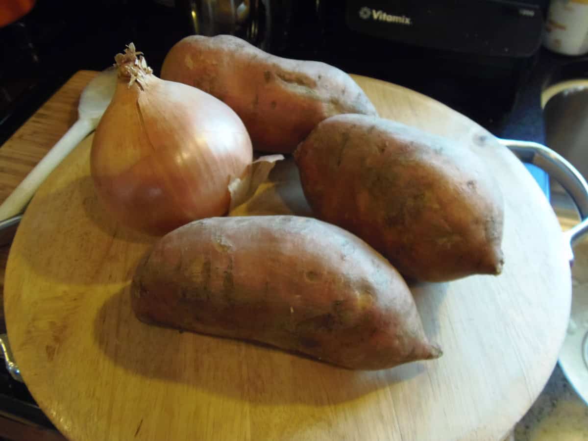 Three large sweet potatoes and one onion on a wooden cutting board.