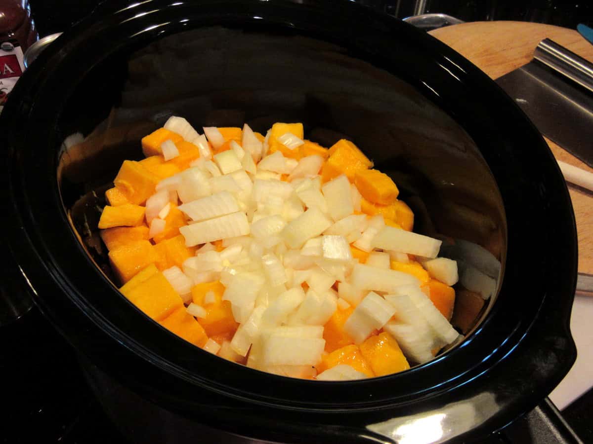 Chopped onions and butternut squash chunks in a black slow cooker insert.