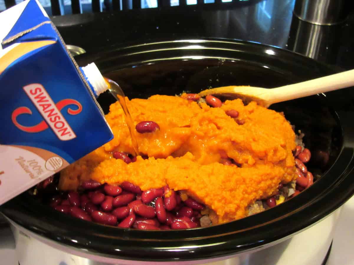 Beef stock being added to a slow cooker that has kidney beans and pumpkin puree inside.