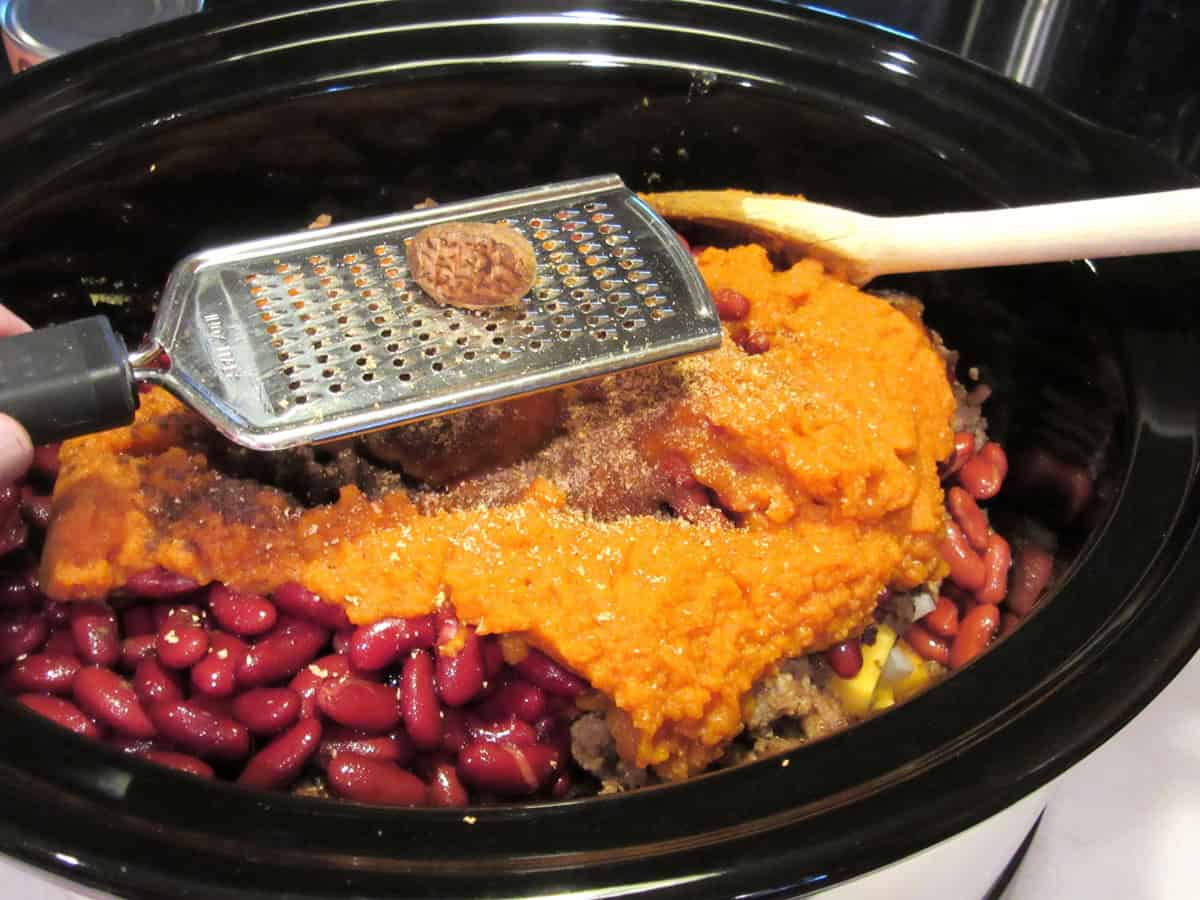 A nutmeg pod being grated into a slow cooker containing the start of butternut squash chili.
