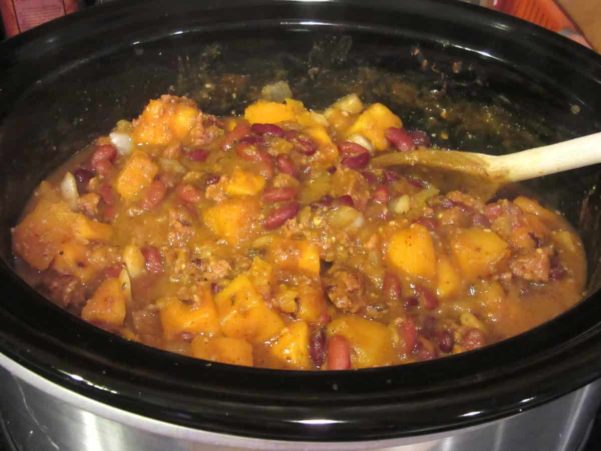 A wooden spoon inside a slow cooker containing butternut squash chili with beans and sausage.