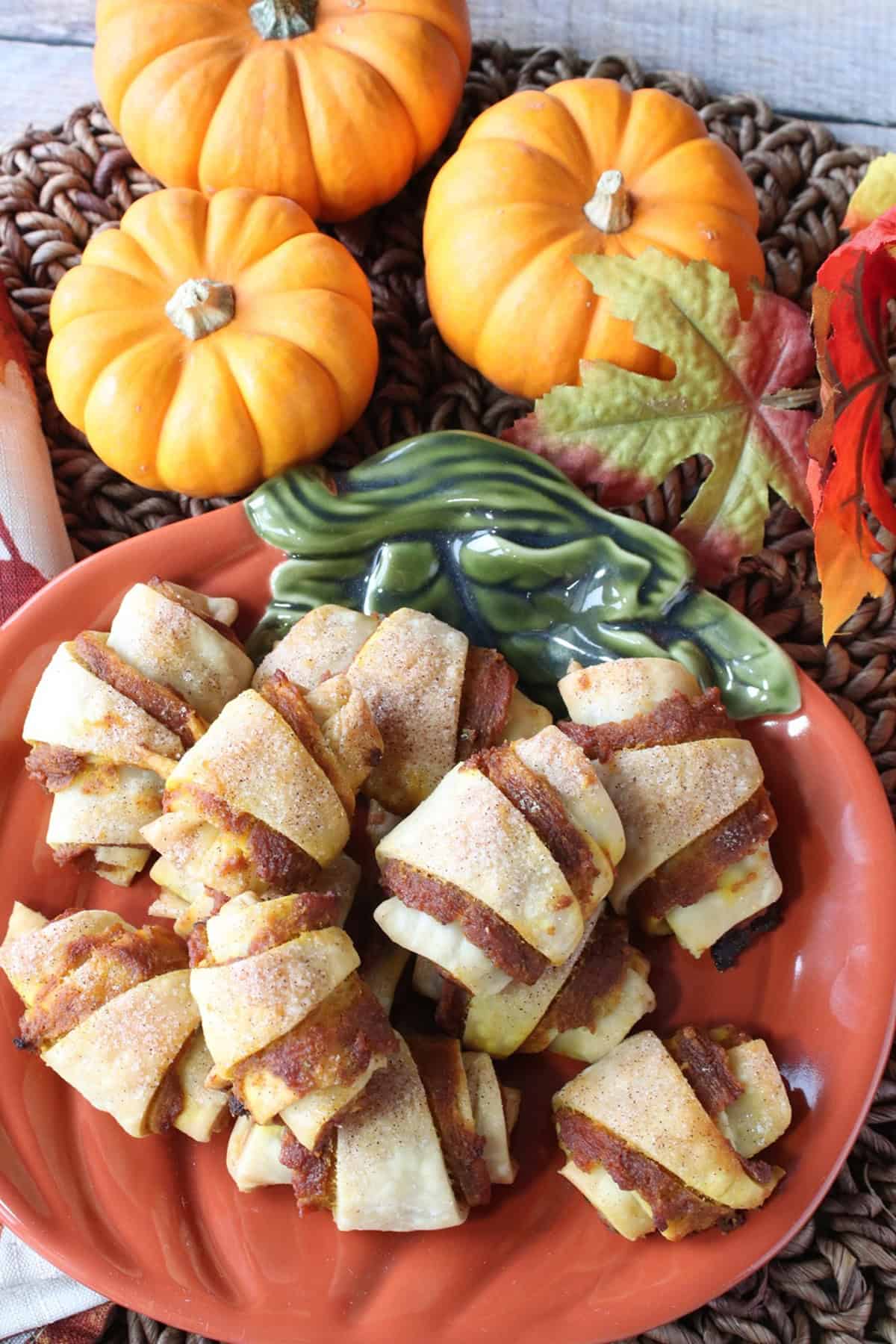Three mini pumpkins in the background of a stack of pumpkin pie bites on a small pumpkin plate.